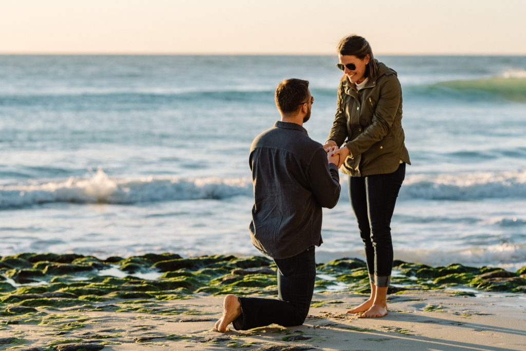 The tide pools at Swami's Beach in Encinitas provide the perfect backdrop for this surprise San Diego Proposal. Man down on one knee - woman holds his hands. 