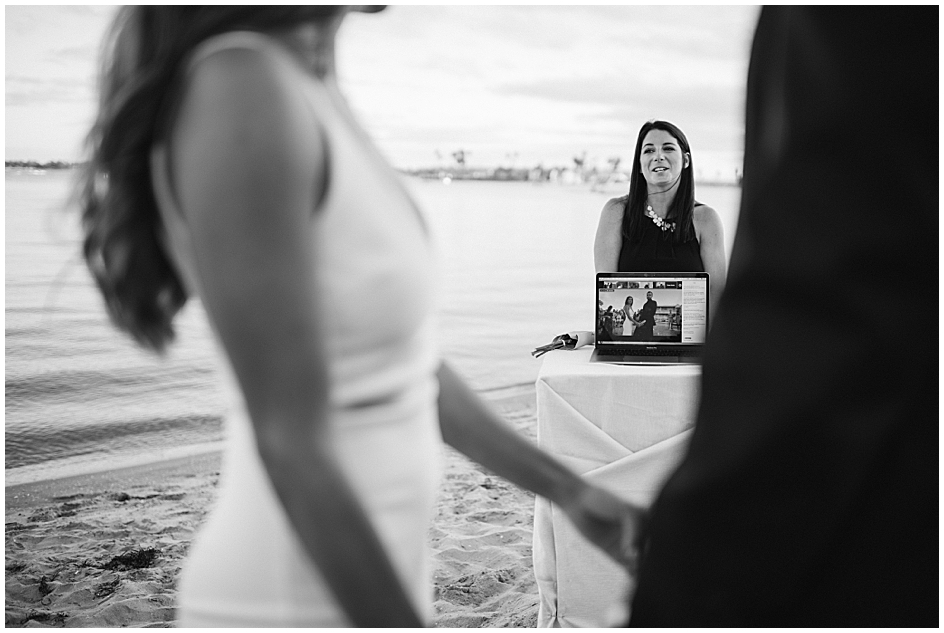 Bride and Groom join hands during their wedding Ceremony in front of a laptop showing their Live-streamed Ceremony to friends and family not in attendance due to the Coronavirus Covid-19 Pandemic at this San Diego Microwedding photographed by Marie Monforte. 