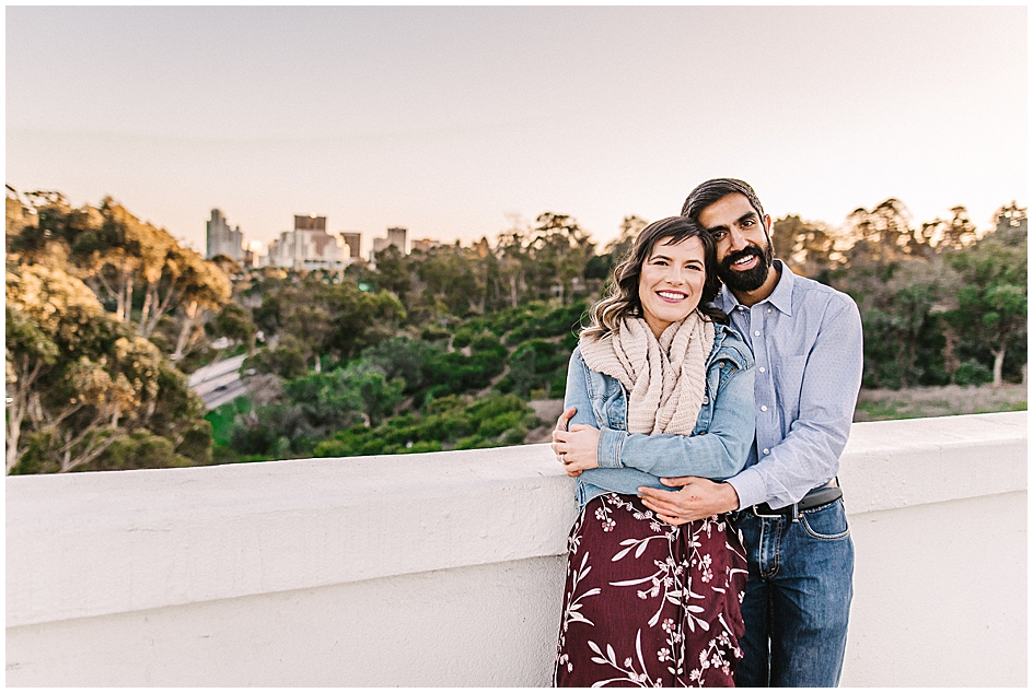 Couple embraces while smiling at the camera on Balboa Park's Cabrillo Bridge - with views of downtown San Diego in the background. 