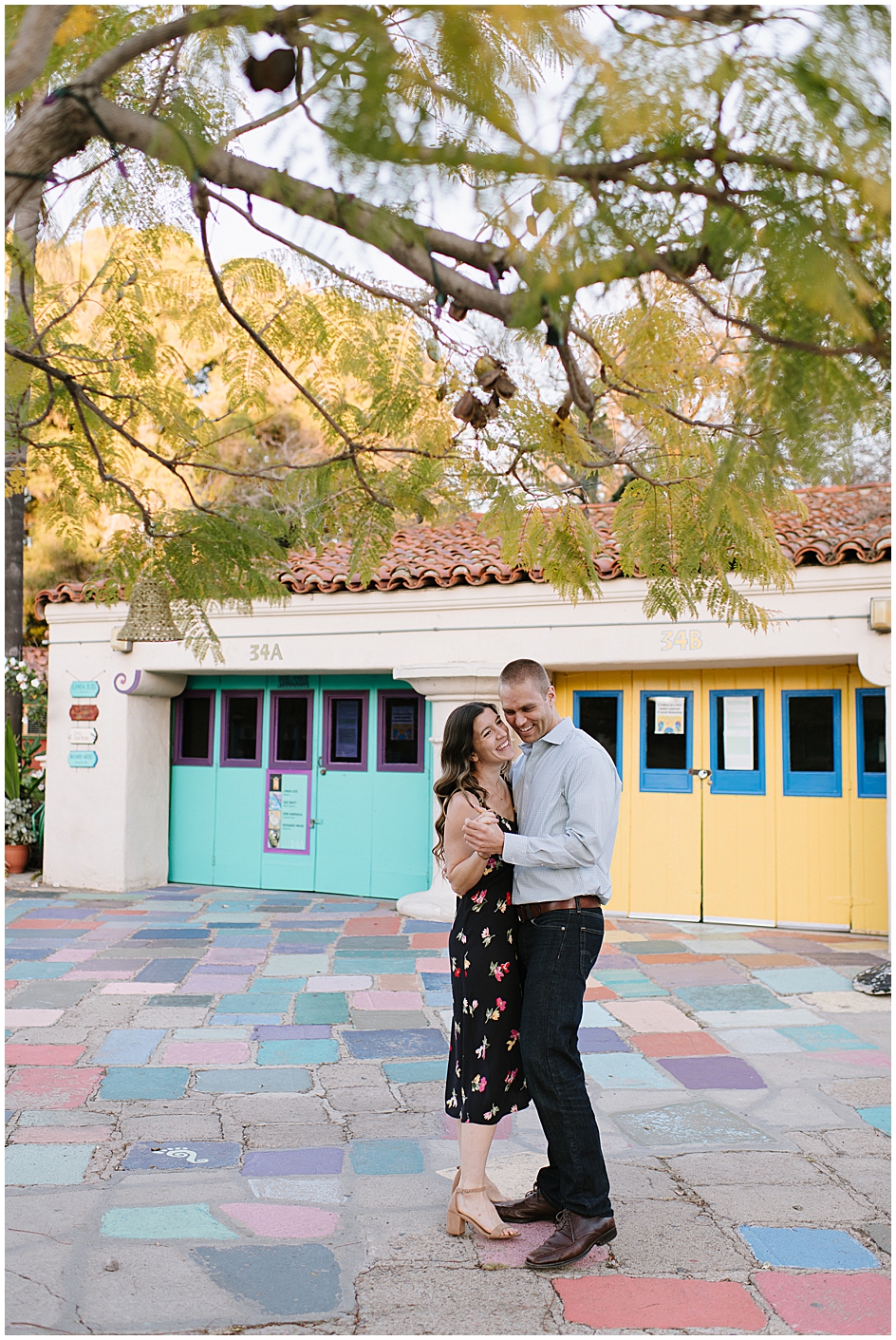 Young couple dancing under the tree in Spanish Village, Balaboa Park. Colorful painted floor stones below them. IN the background artist studios with yellow and turquoise doors.