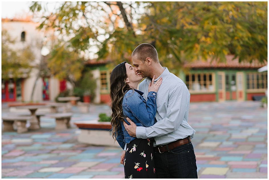 Young couple shares a kiss during their engagement photos in Balboa Park's Spanish Village.  The whimsical and colorful background of painted stone floor tiles and artist studios is one of Balboa park's best spots for engagement photos. 