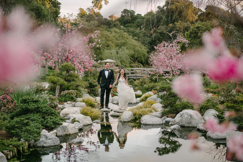 Bride and Groom stand on rocks in the middle of the stream, surrounded by pink cheery blossoms in Balboa's park's Japanese Friendship Garden. Captured by San Diego Wedding Photographer Marie Monforte. 