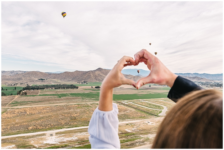 Recently engaged couple press their two hands together to form a heart framing a Hot Air Balloon in the distance.  
