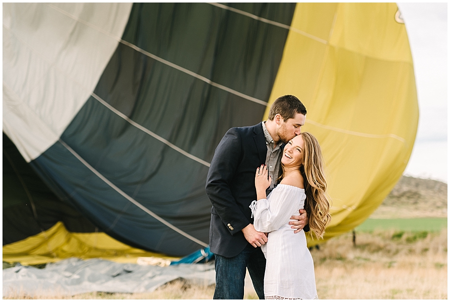 Man gives his new fiancee a kiss on the temple while they hug and she laughs off to the side as the Hot Air Balloon behind them deflates during this Temecula Engagement Session. 