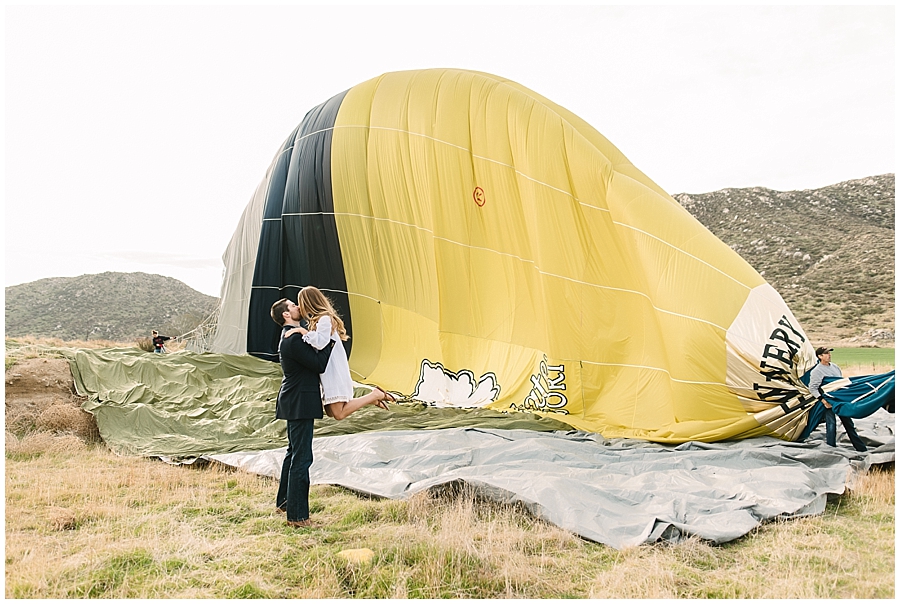 Man sweeps his new fiancee off her feet against a backdrop of a Hot Air Balloon in this Temecula engagement session by San Diego Photographer Marie Monforte. 