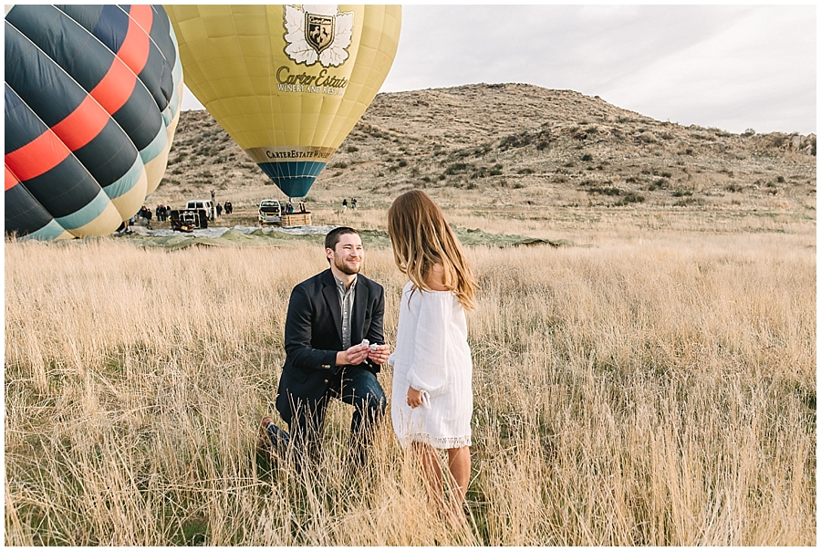 Early sunrise in a Temecula field of golden grasses, a man in a dark suit bends his knee to propose to his girlfriend, smiling wide. Behind him hot air balloons are filling up in the distance as they are about to embark on a hot air balloon ride.
