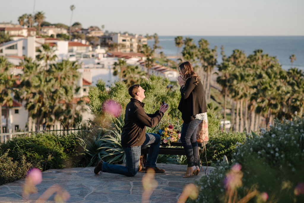 Man down on one knee for his San Diego Proposal Session overlooking the ocean, palm trees, and a stretch of spanish style condominiums. 