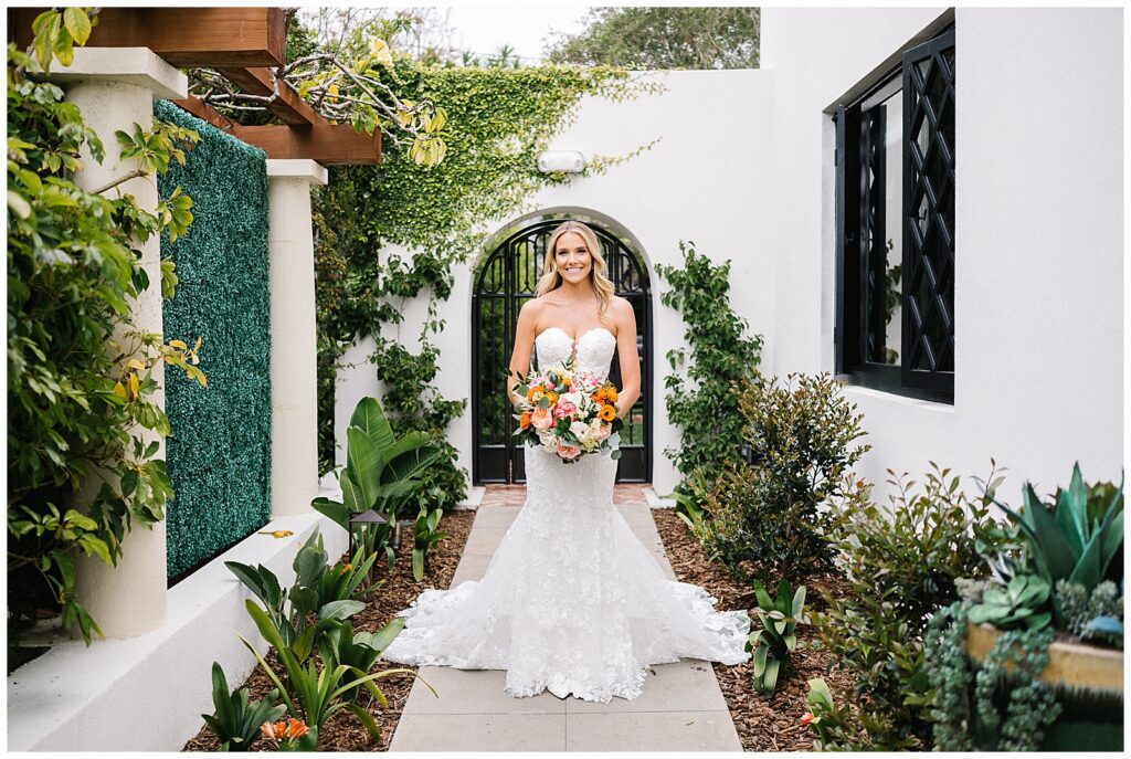 A smiling bride in a lace white dress holds a colorful wedding bouquet and walks along a bright, beautiful courtyard. 