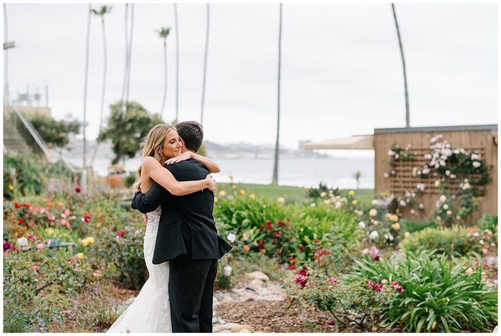 A bride and groom embrace each other in a colorful garden next to the ocean before their wedding ceremony. 