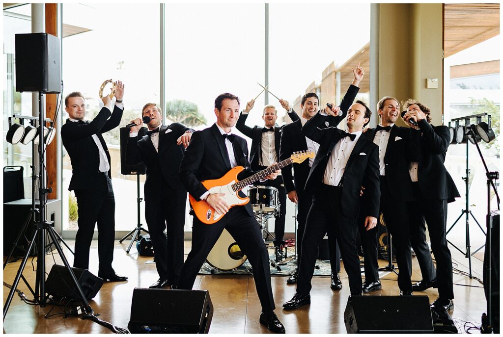 Men in black suits and black bowties all smile as they play instruments in  front of a big window on a stage. 