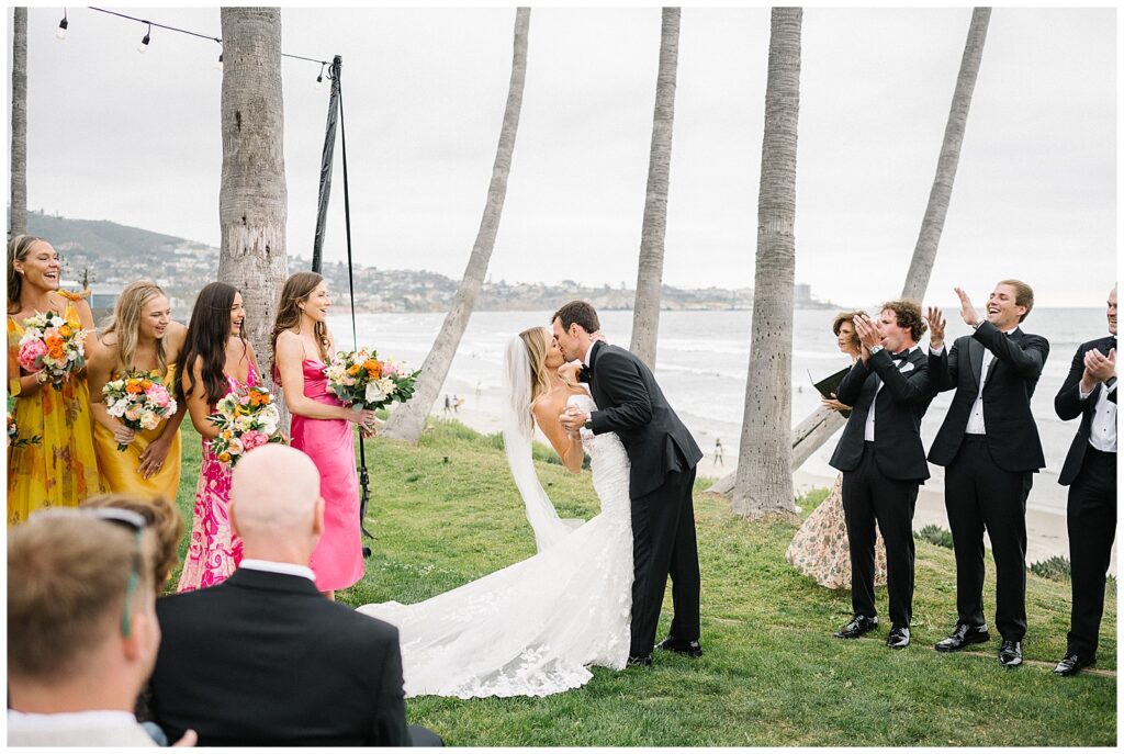 On top of a cliff next to the ocean in San Diego, a bride and groom hold hands and kiss during their wedding ceremony as their family and friends clap and cheer. 