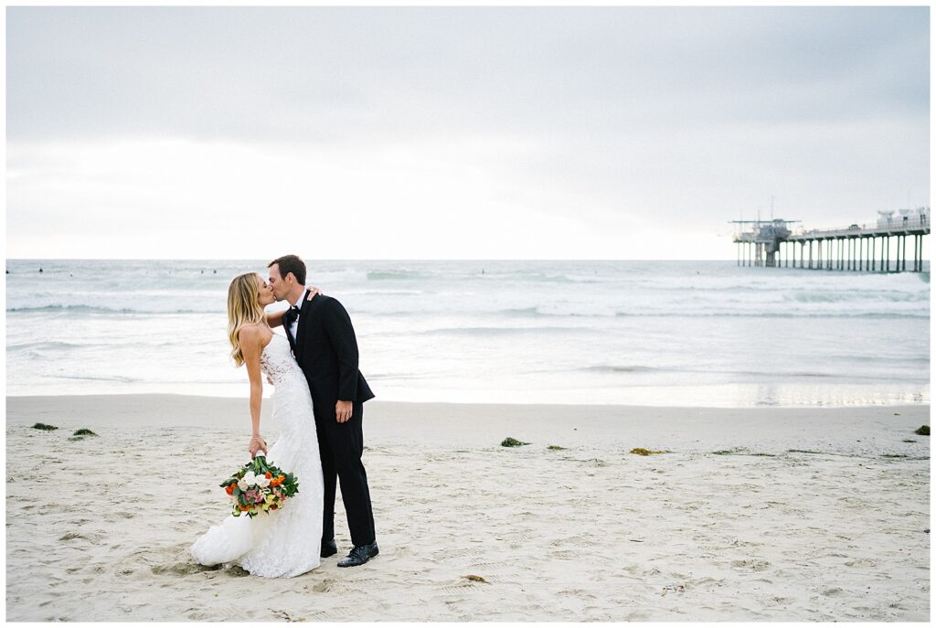 On an overcast beach next to a pier, a bride holds a colorful bouquet of flowers and kisses her husband after their wedding ceremony. 