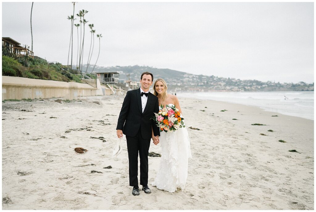 A just-married couple holds hands and smiles as they walk down a cloudy beach in San Diego. 