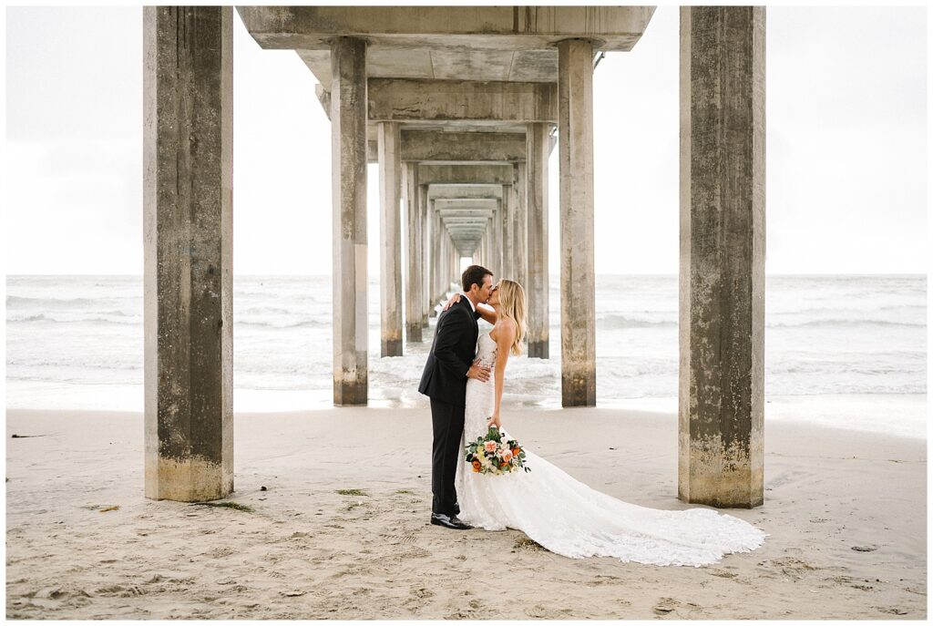 Dressed in their wedding attire, a bride and groom share a kiss under a stone pier in San Diego, California. 
