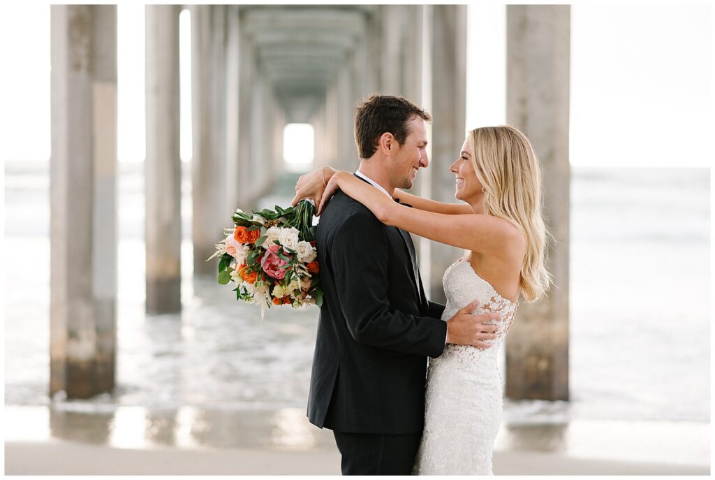 A bride and groom embrace each other and laugh under a stone pier in San Diego, California after their wedding ceremony. 